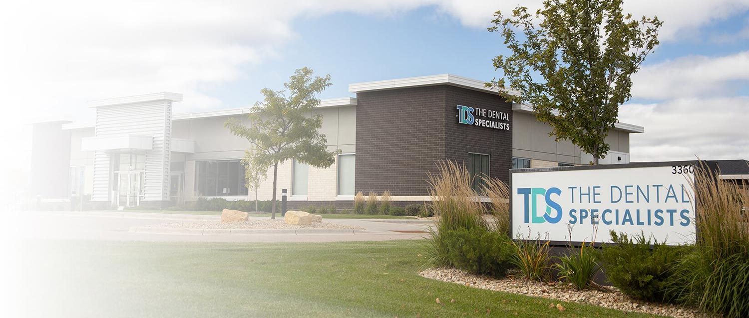 The Dental Specialists practice exterior with a sign displaying the logo and name, surrounded by landscaping and trees.